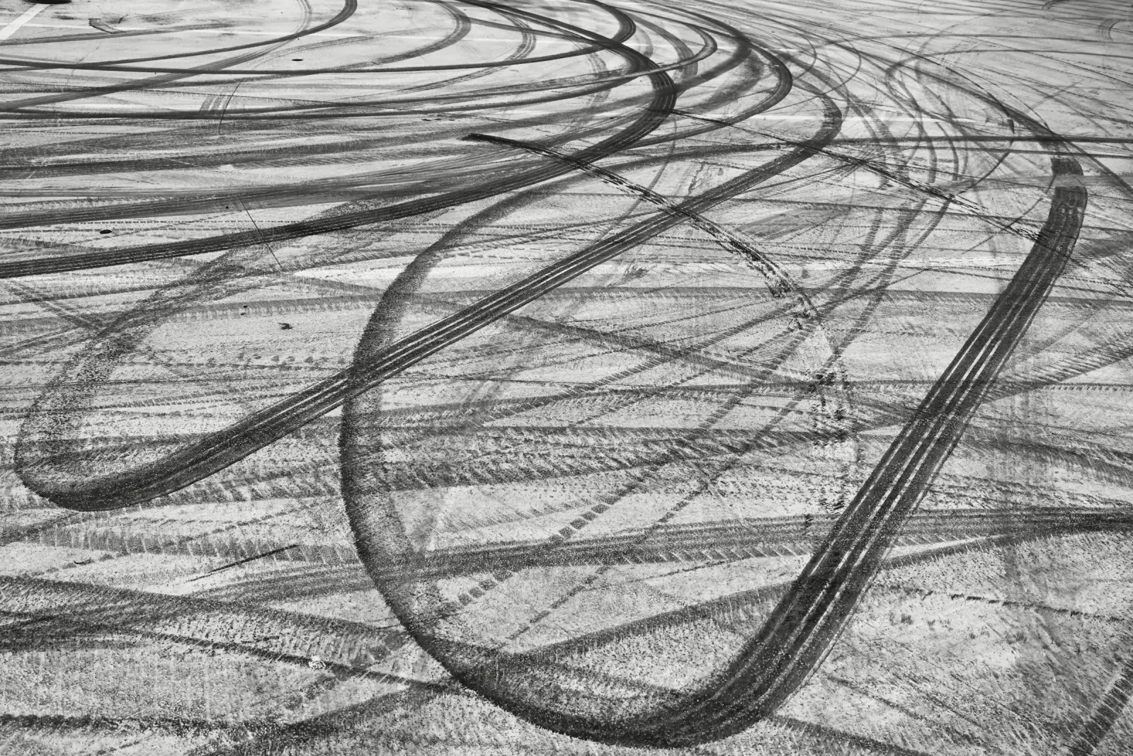 Black-and-white photograph of overlapping tire tracks curving across pale pavement, layered marks left by many passes.
