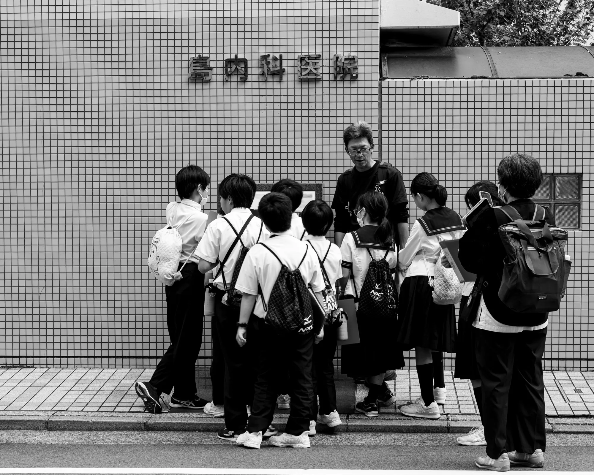 A teacher in Japan speaking with a group of schoolchildren wearing backpacks, gathered outside against a wire-mesh fence.